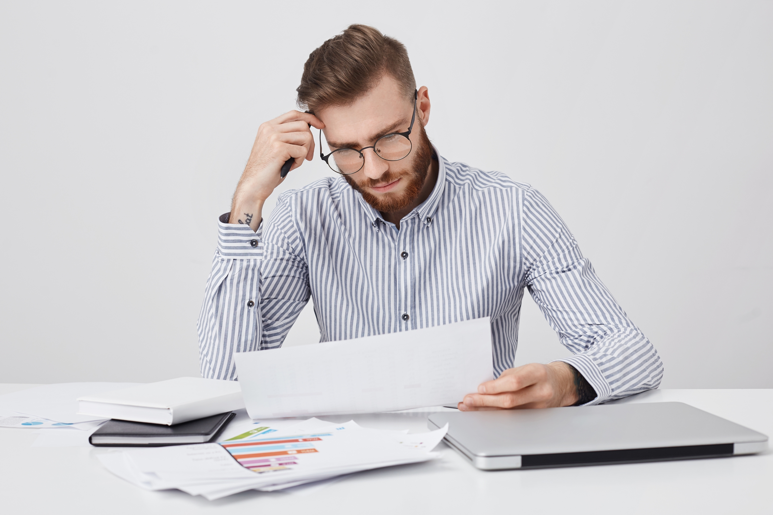 Serious concentrated bearded man dressed formally, reads document or contract, sits at work place, tries to understand figures and diagrams. Successful male enterpreneur studies office papers alone